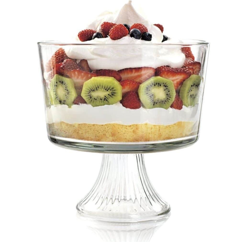 A layered dessert in a glass stand bowl. This easy fruit trifle recipe begins with a yellow cake base, topped with a white creamy layer, then sliced kiwis, strawberries, raspberries, and blueberries. It's finished with whipped cream and more berries on top.